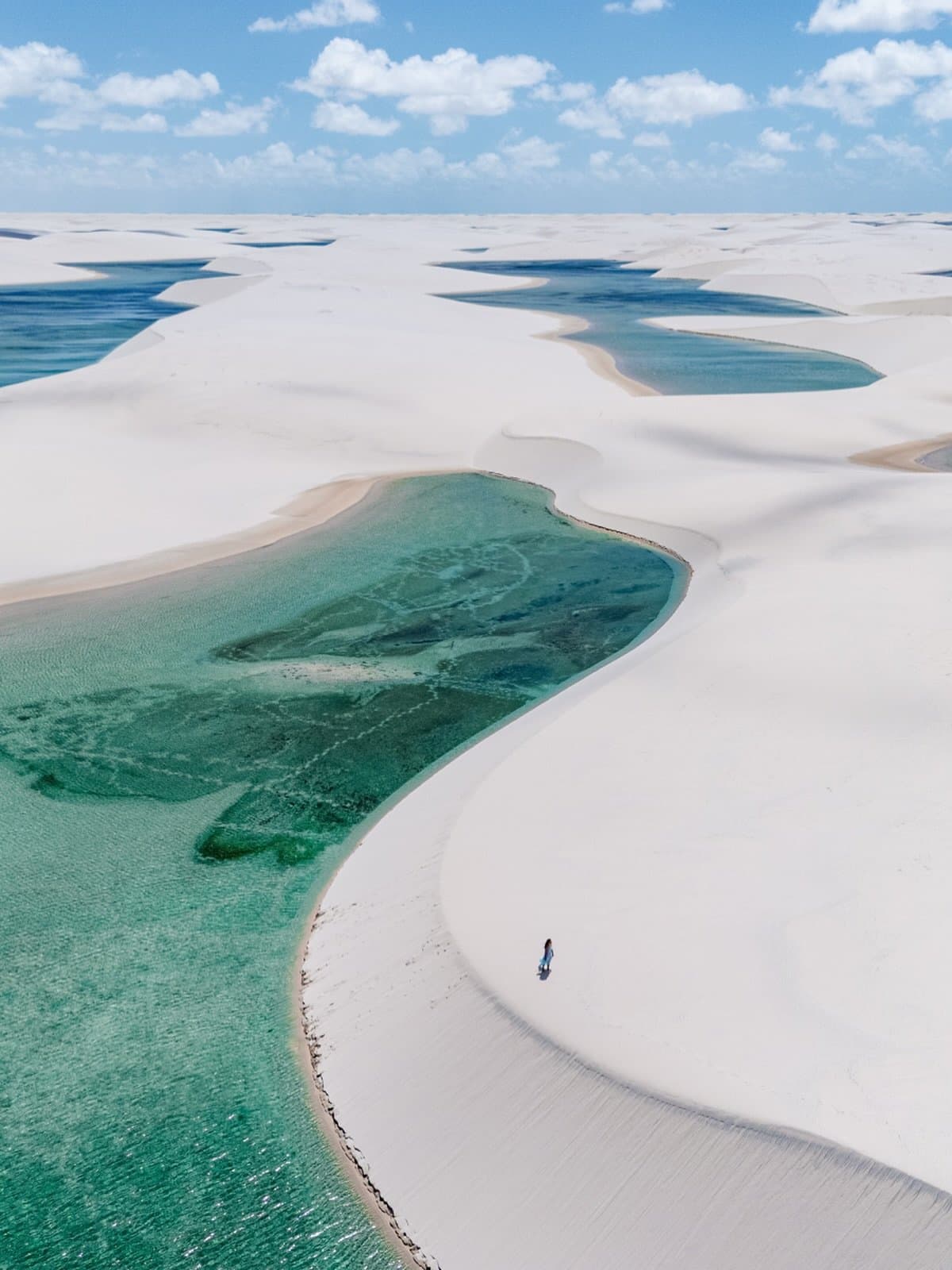 Un désert qui se noie dans l'eau douce, et on ne s'en plaint pas. Vue aérienne des Lençóis Maranhenses près de Santo Amaro, montrant de vastes dunes de sable blanc parsemées de lagunes turquoise et émeraude s'étendant jusqu'à l'horizon, avec une seule personne au bord de l'eau sous un ciel bleu