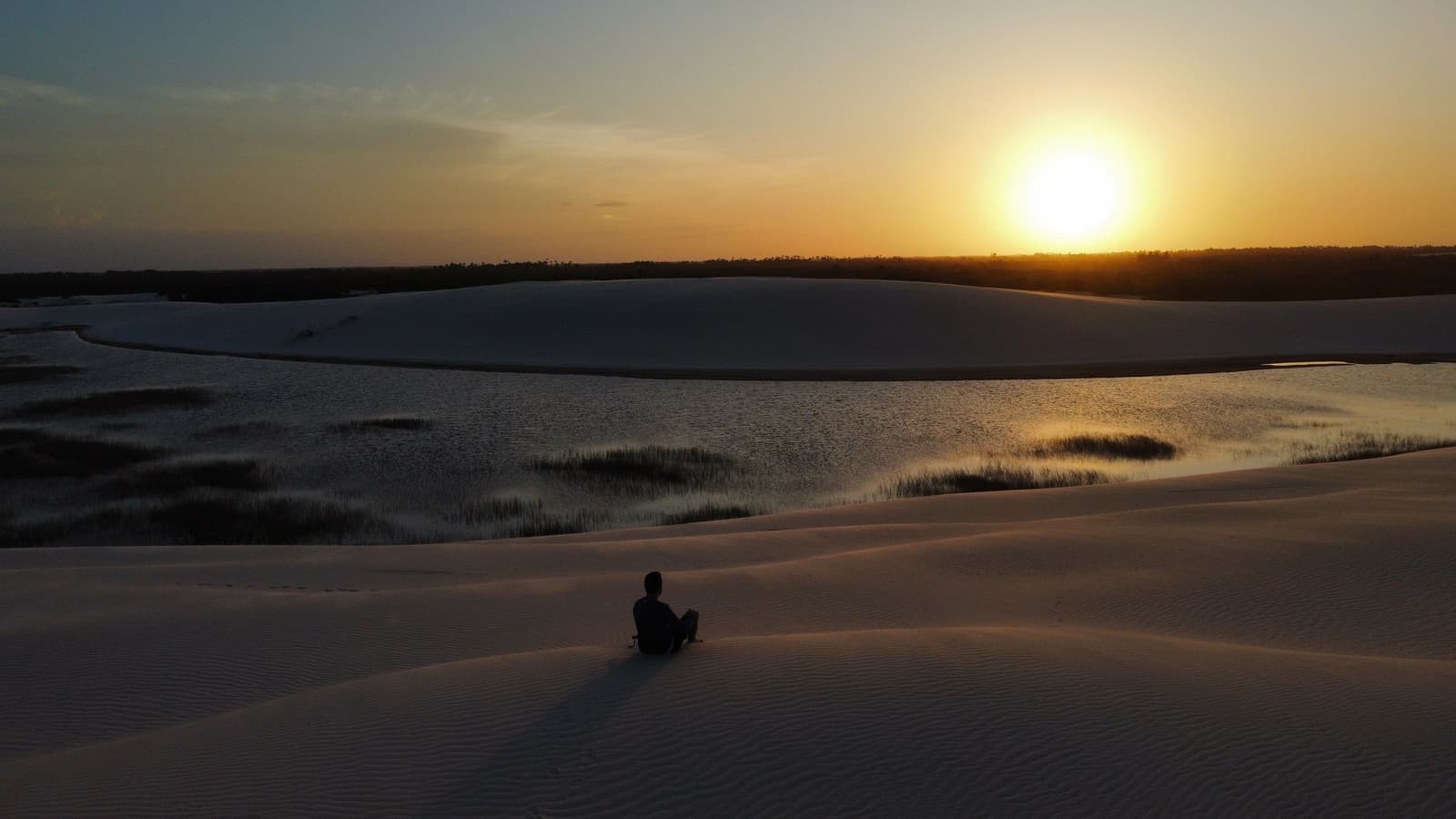 Certains couchers de soleil méritent qu'on s'arrête et qu'on ne dise rien. Une personne assise seule sur une dune de sable blanc à Lençóis Maranhenses près de Santo Amaro, regardant le soleil se coucher sur un lagon calme bordé d'herbes et d'une ligne d'arbres lointaine