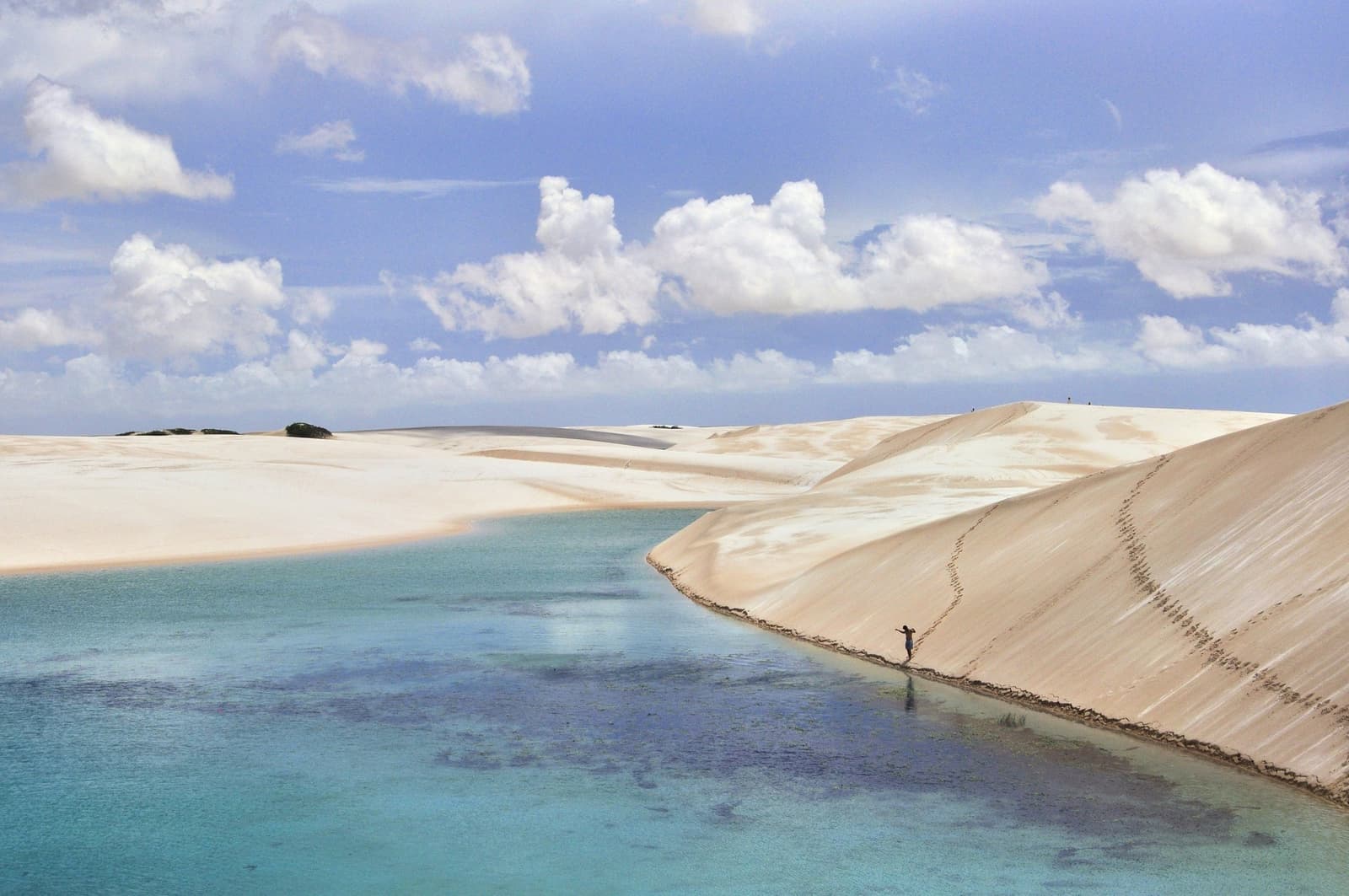 Uma lagoa, uma pessoa, e dunas que fazem o mundo parecer maior. Um caminhante solitário percorre a borda de uma duna de areia branca ao lado de uma lagoa turquesa durante o trekking nos Lençóis Maranhenses saindo de Atins, com nuvens refletidas na água calma