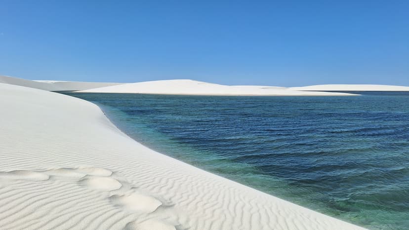 Rippled white sand dunes meeting a wide turquoise-blue freshwater lagoon under a clear blue sky on the Lençóis Maranhenses trek from Atins Rippled white sand dunes meeting a wide turquoise-blue freshwater lagoon under a clear blue sky on the Lençóis Maranhenses trek from Atins