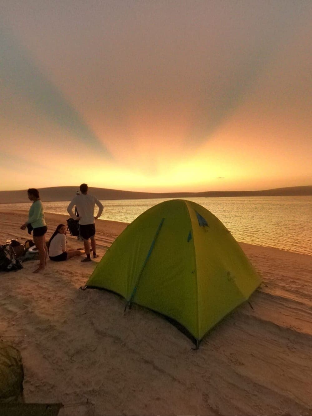 Ninguém precisou combinar parar, aconteceu naturalmente. Barraca dôme verde armada numa praia de areia durante o trek pelos Lençóis Maranhenses, com três viajantes observando um pôr do sol dourado sobre água calma e dunas ao fundo
