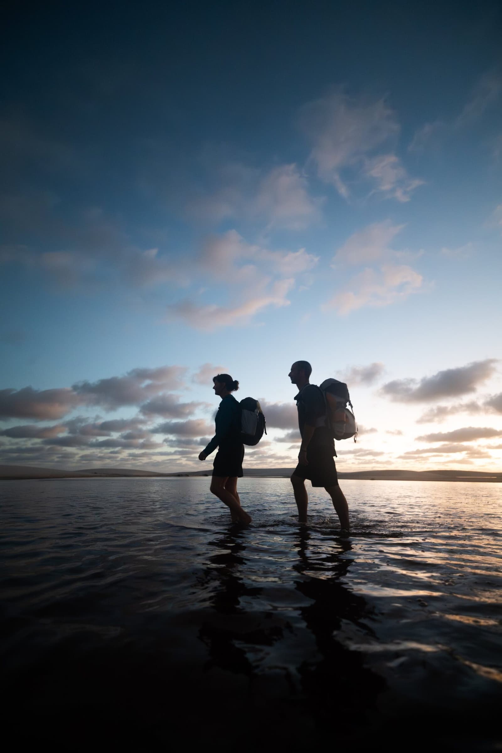 Pés molhados, mochila nas costas, céu que vale cada passo. Duas pessoas com mochilas em silhueta contra o céu ao entardecer, atravessando descalças uma lâmina d'água rasa durante o trek pelos Lençóis Maranhenses