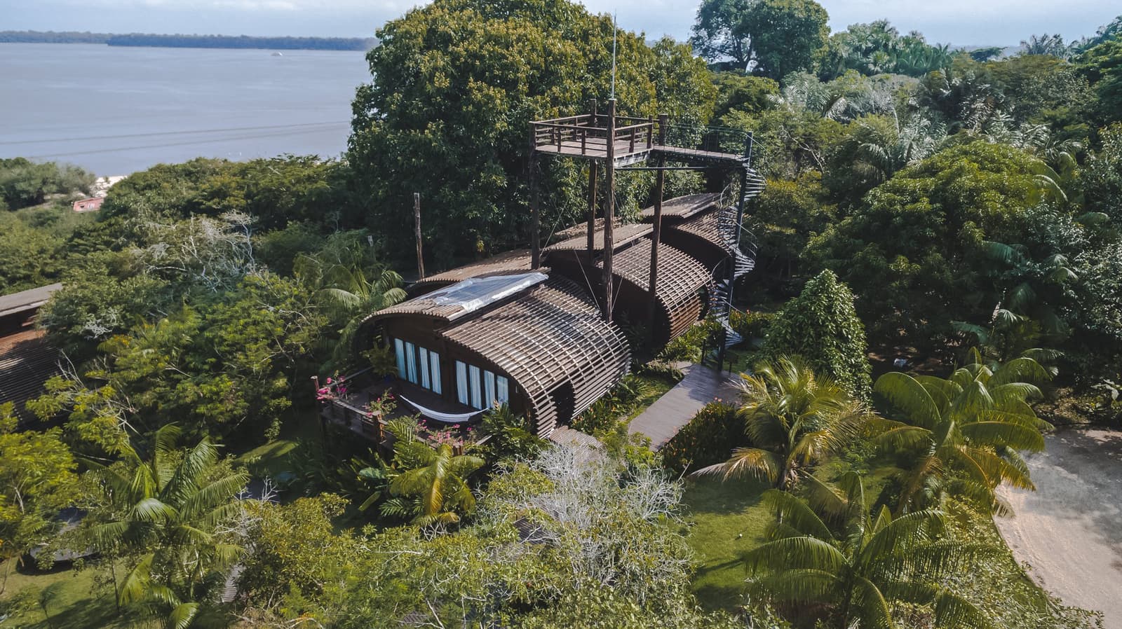 On dirait que la forêt a décidé de construire son propre hôtel. Vue aérienne du lodge Mirante do Gavião avec sa structure en lattes de bois courbées, sa passerelle dans la canopée et sa tour d'observation dominant la forêt tropicale au bord d'un large fleuve