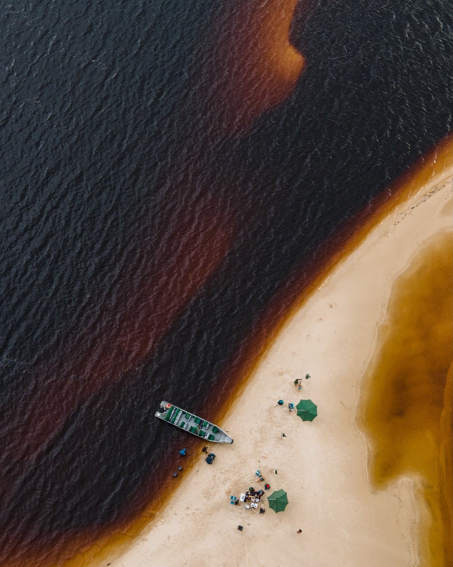 Deux rivières, un banc de sable, et un bateau qui a visiblement fait le bon choix. Vue aérienne d'une petite plage de sable sur le Rio Negro près de Mirante do Gavião, où les eaux noires rencontrent un rivage aux reflets ambrés, avec une barque amarrée au bord de l'eau et deux parasols verts abritant des visiteurs