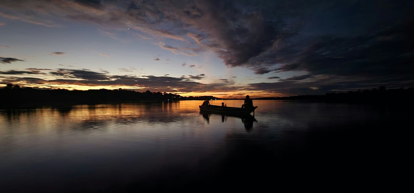 Le fleuve garde le ciel pour lui, juste le temps du crépuscule. Silhouette de trois personnes dans une petite embarcation en bois sur une eau calme au crépuscule, le coucher de soleil du Mirante do Gavião teintant l'horizon d'or et de violet sous des nuages dramatiques