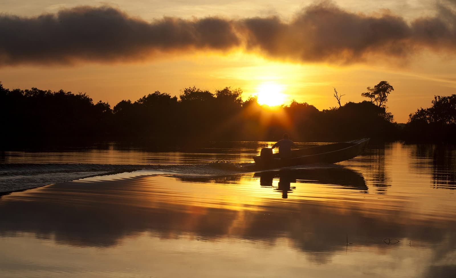 Le fleuve ne se presse jamais, et on finit par lui donner raison. Une silhouette pilote un hors-bord sur un fleuve lisse comme un miroir au coucher du soleil doré près du Mirante do Gavião, le soleil perçant les nuages au-dessus d'une ligne d'arbres sombre