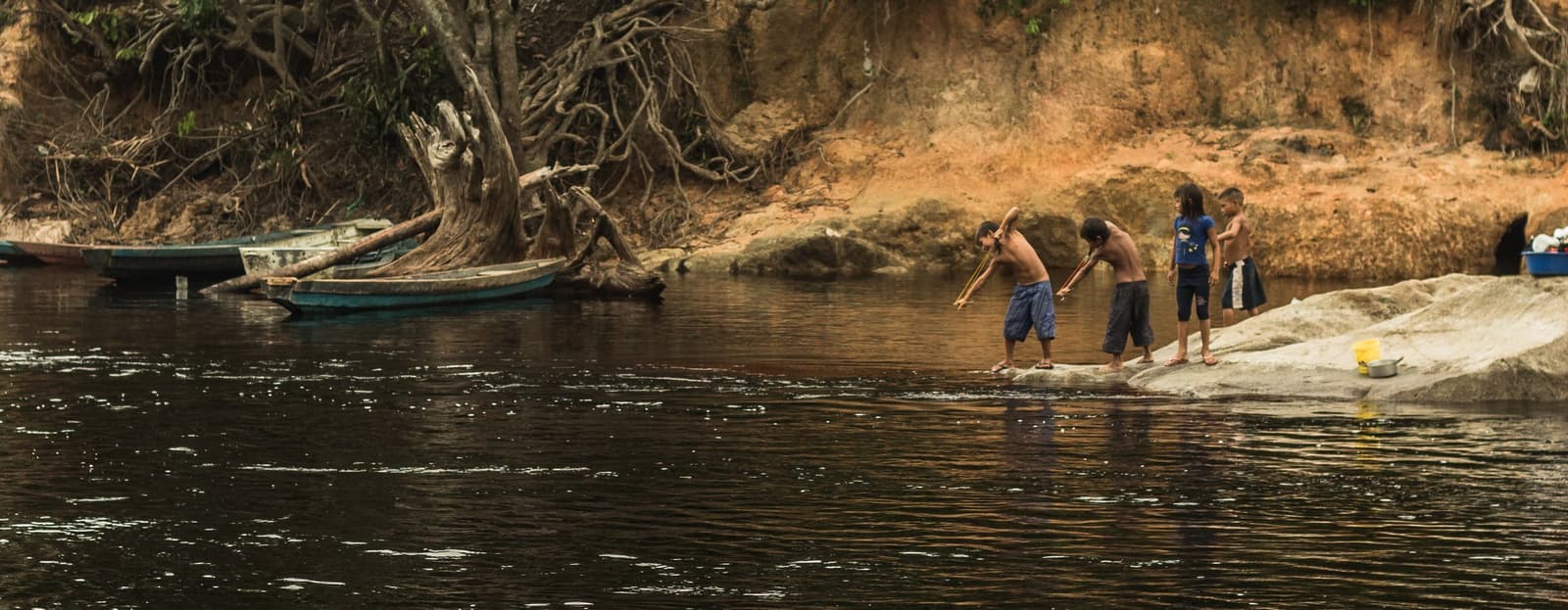 Vier kinderen, lange stokken en een rivier die geen uitleg nodig heeft. Vier kinderen staan op een vlakke rots aan de oever van een donkere Amazonerivier en vissen of spelen met lange stokken, met houten kano's en blootliggende boomwortels langs een zandige kleiwal op de achtergrond