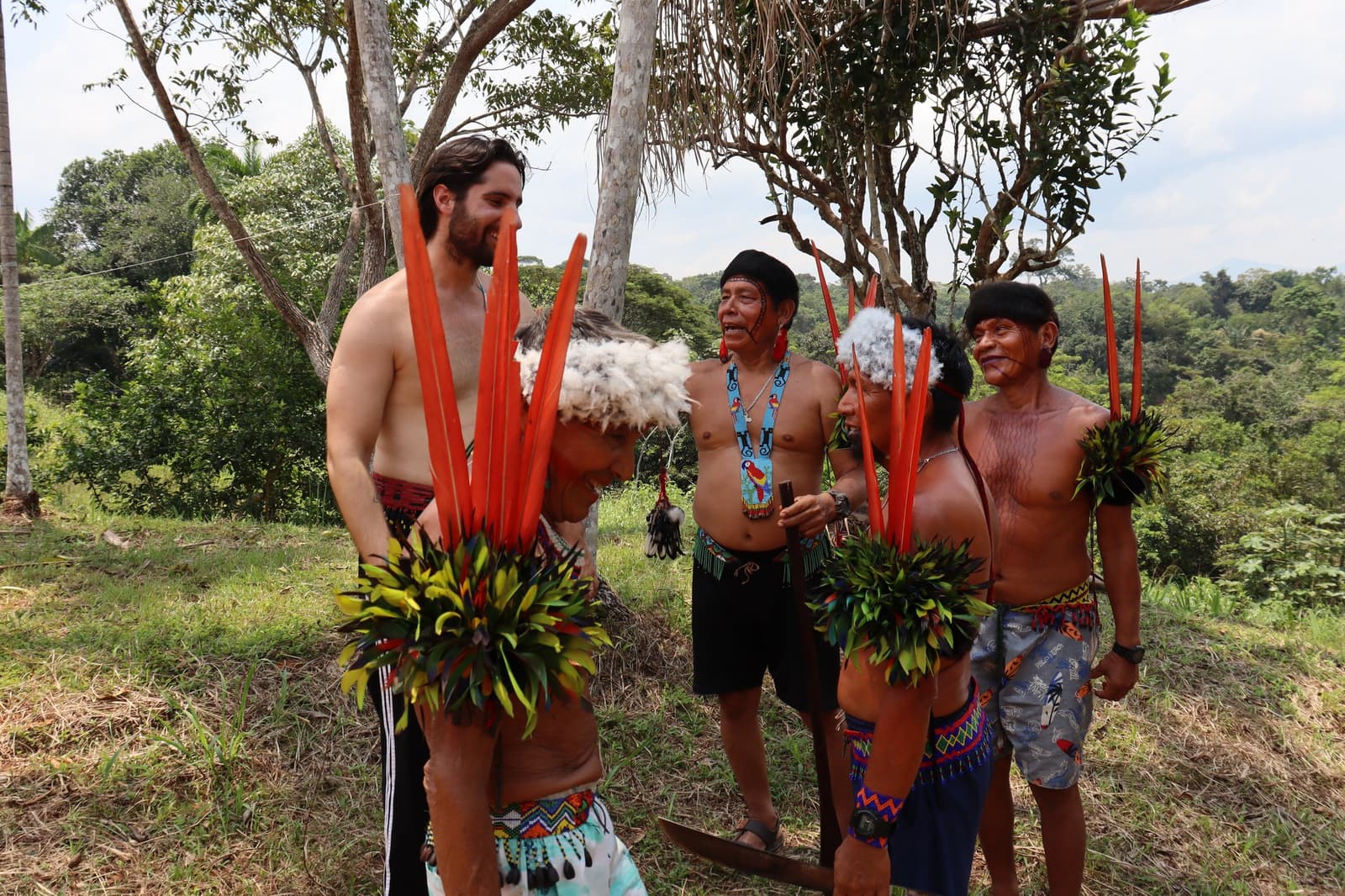 Aangekleed door de gemeenschap, samen lachend. Sommige uitnodigingen accepteer je gewoon. Een lachende westerse reiziger met een traditionele vedertooi met lange rode veren staat tussen Yanomami-mannen tijdens een culturele ontmoeting op de Pico da Neblina Expeditie, omgeven door Amazoneregenwoud
