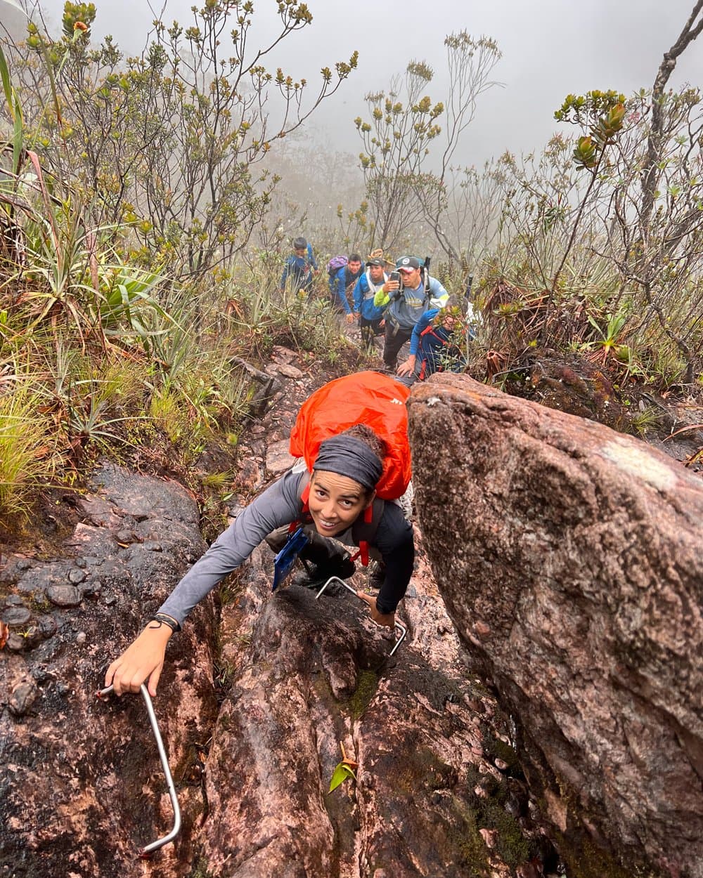 Handen aan de rots, rugzak op de rug, glimlach onveranderd. Een glimlachende vrouwelijke trekker met een rode regenhoes op haar rugzak klimt over een natte, met mos bedekte rotswand tijdens de Pico da Neblina Expeditie, terwijl ze een bevestigd metalen handvat vastgrijpt en een rij trekkers in de mist achter haar volgt