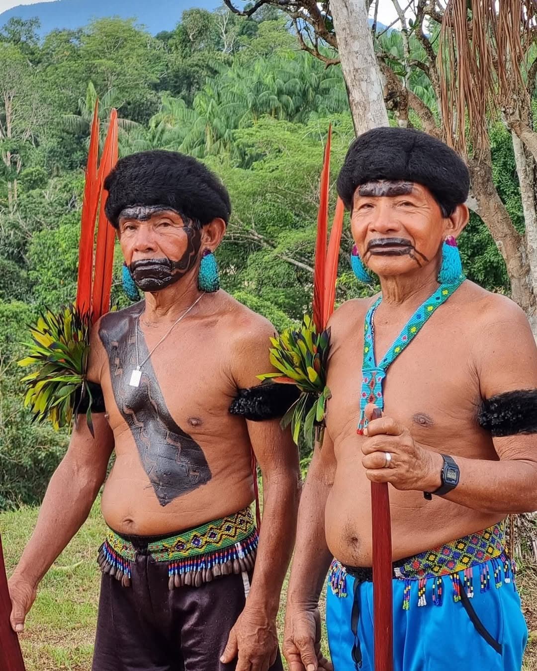 Voor de eerste stap krijgt het woud het woord. Twee oudere Yanomami-mannen in traditionele ceremoniële kleding — zwarte lichaamsverf, rode verentooi, turquoise kraaloorbellen en geweven kraalgordels — staand voor het regenwoud