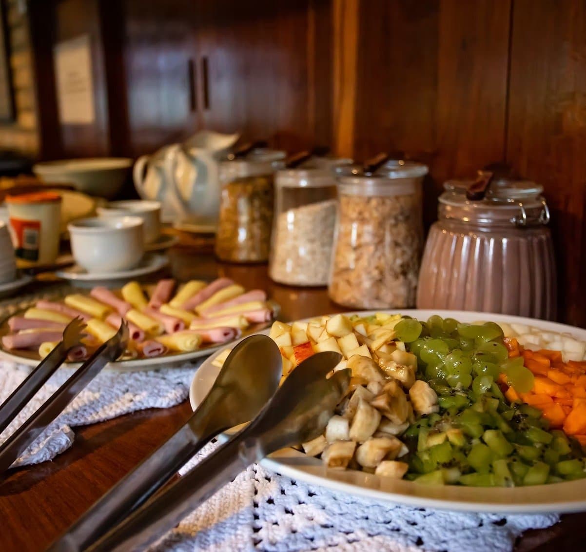 On part mieux dans la nature quand le ventre est plein. Buffet petit-déjeuner d'un lodge de safari dans le sud du Pantanal avec des fruits frais tranchés comme banane, kiwi, raisin et papaye, des charcuteries roulées et du fromage, des bocaux de granola, des tasses en céramique et une cruche sur un buffet en bois sombre
