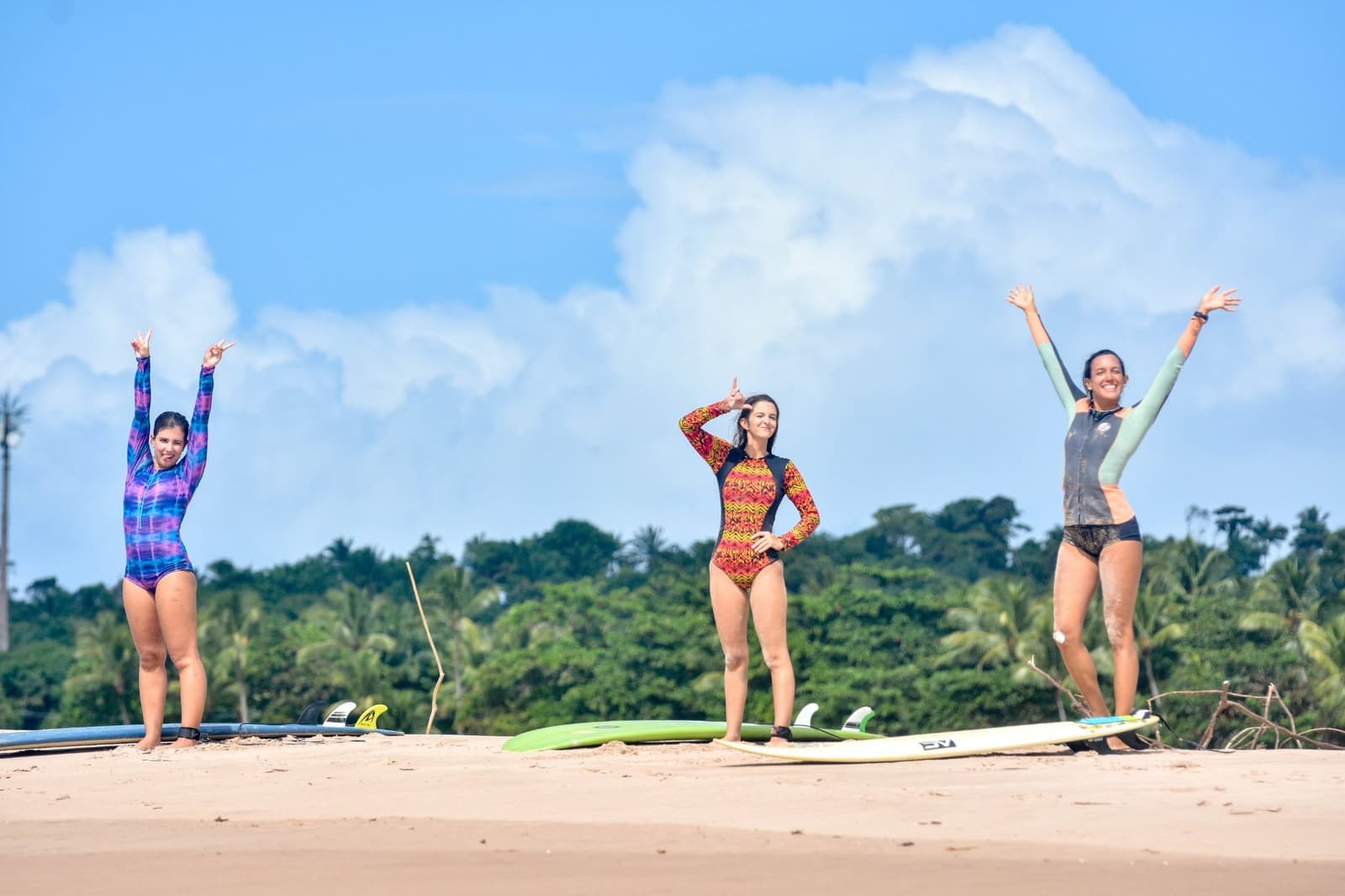 Als je les krijgt met dit uitzicht, vergeet je dat het ook gewoon hard werken is. Photo: Picasa Drie vrouwen in kleurrijke rash guards vieren feest op een zandstrand met hun surfboards tijdens surfles in Itacaré, met een weelderige groene bomenrij en blauwe lucht op de achtergrond.