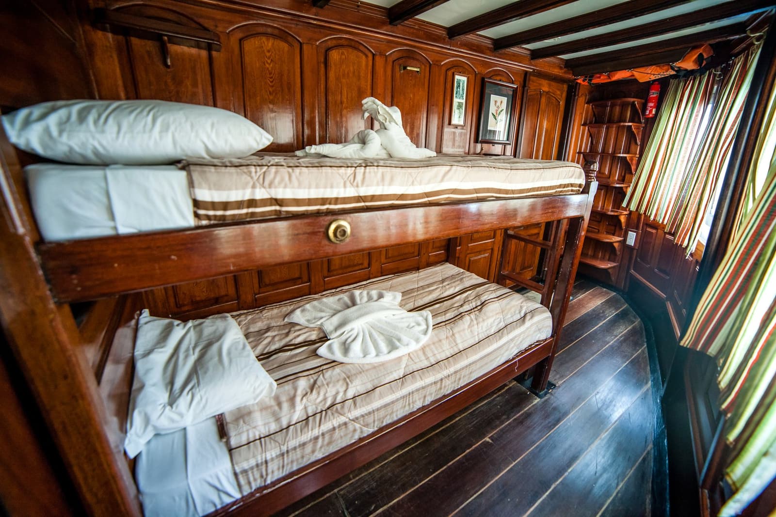Knus en compact, met handdoekkunst als welkomstgroet. Photo: er: Houten stapelbedkabine aan boord van de Tucano Amazon-riviercruise, met gestreept beddengoed, in dierenvorm gevouwen handdoeken en gelakt houten betimmeringswerk