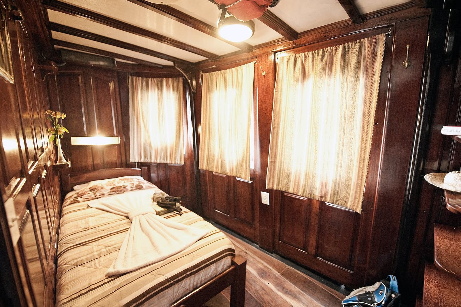 Niet groot, maar na een dag in de jungle voelt het als een paleis. Sfeervolle kajuitinterieur van de Tucano Amazone riviercruise met donker gelakt houtpaneel, een tweepersoonsbed met gestreept beddengoed, warm wandlicht, goudkleurige gordijnen en een klein vaasje met gele bloemen