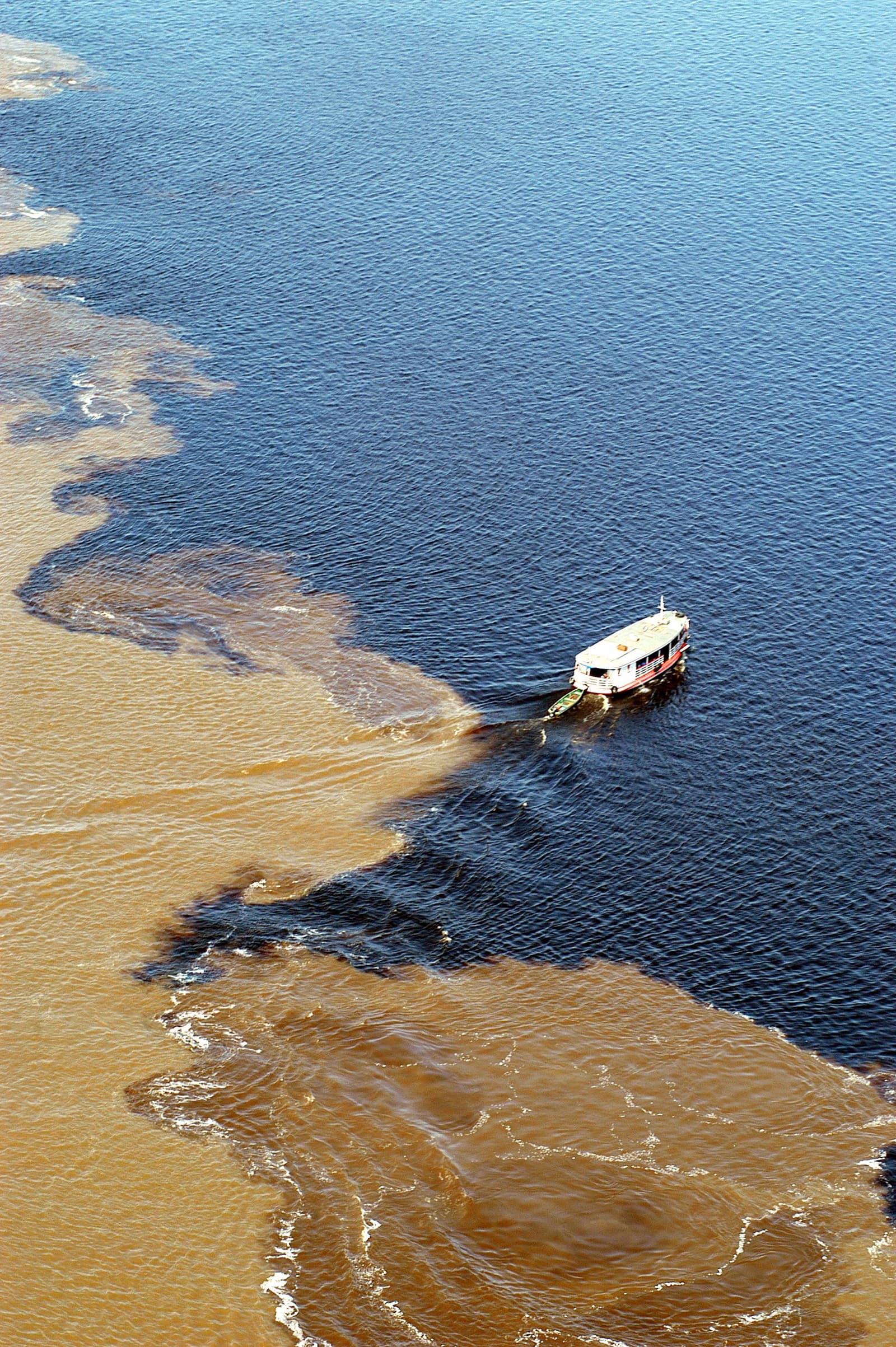 Twee rivieren, één lijn ertussen, geen akkoord. Luchtfoto van een riviercruiseschip dat de Ontmoeting der Wateren oversteekt, waar donker zwart water en zandbruin, sedimentrijk water naast elkaar stromen zonder te vermengen