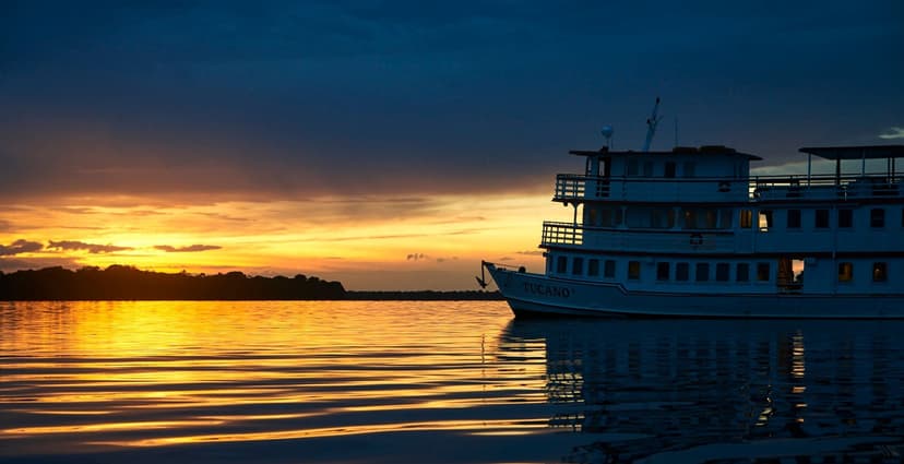 The M/Y Tucano, a white multi-deck river cruise vessel, moored on a calm river at sunset, with a vivid orange and gold sky reflected on the dark water and a treeline silhouetted on the horizon The M/Y Tucano, a white multi-deck river cruise vessel, moored on a calm river at sunset, with a vivid orange and gold sky reflected on the dark water and a treeline silhouetted on the horizon