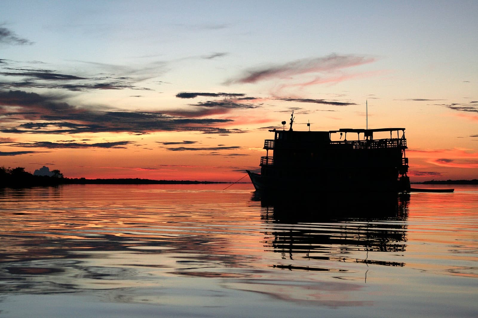 De zon gaat onder, het schip blijft stil, de rivier niet. Een meerdecks rivierschip als silhouet tegen een levendig oranje en rood avondrood dat weerspiegelt in het kalme donkere water, gezien tijdens de Tucano Amazon rivier cruise