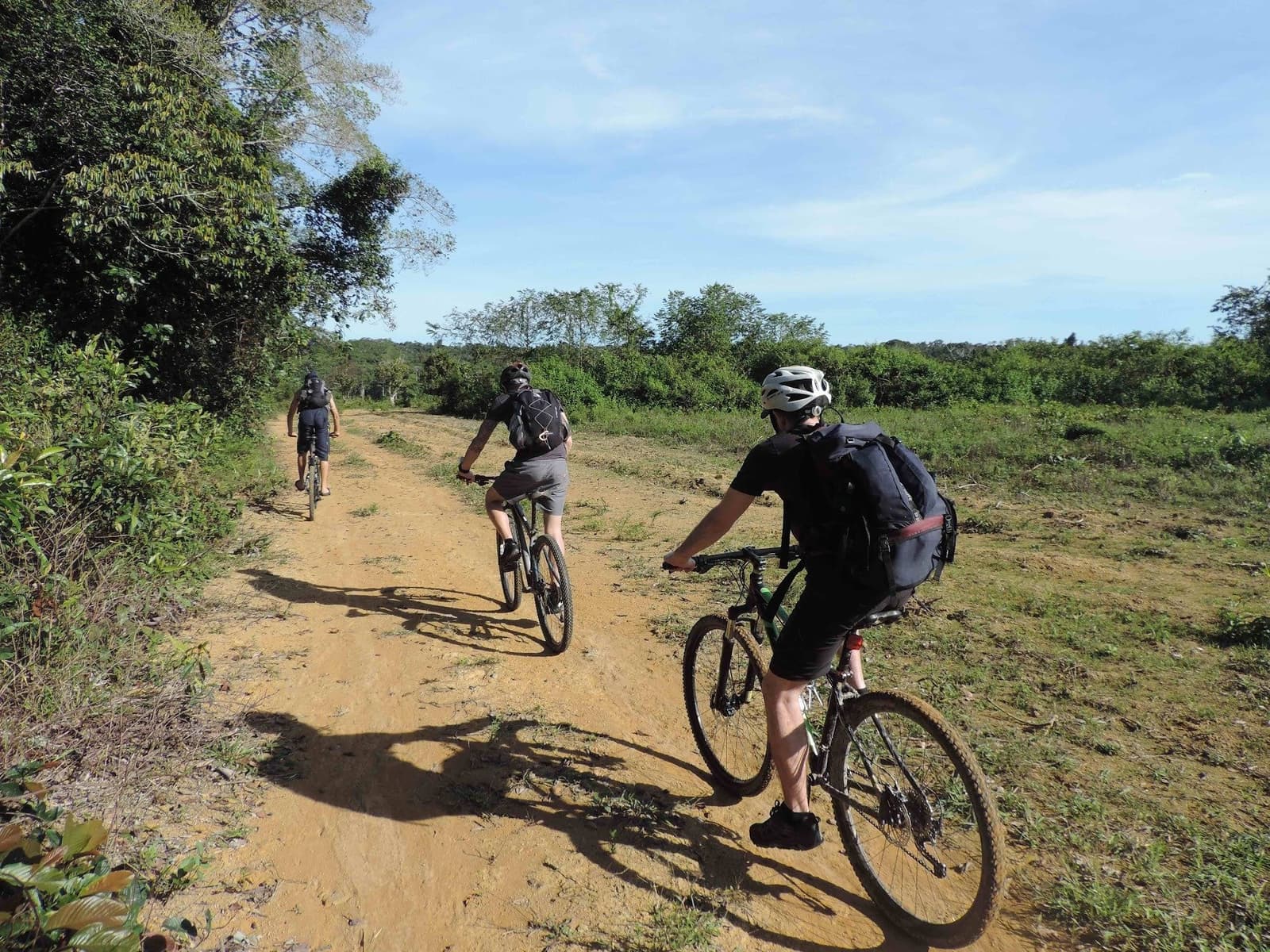 La piste rouge, le vert dense, et personne pour demander si on est arrivés. Trois cyclistes casqués et munis de sacs à dos roulent en file indienne sur une piste de terre rouge lors d'une aventure dans la jungle amazonienne au Brésil, entourés d'une végétation luxuriante sous un ciel bleu