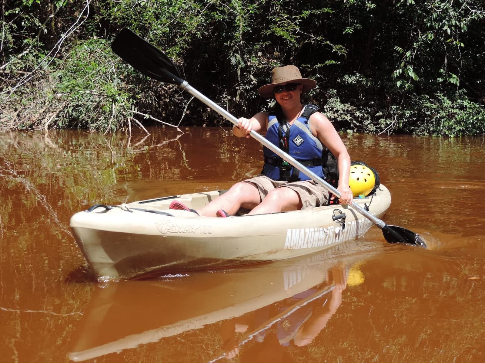 La forêt à portée de pagaie, le sourire en bonus. Une femme souriante coiffée d'un chapeau de soleil et portant un gilet de sauvetage pagaie un kayak solo sur un canal étroit aux eaux brunes tanniques lors d'une aventure dans la jungle amazonienne au Brésil, avec une végétation dense des deux côtés