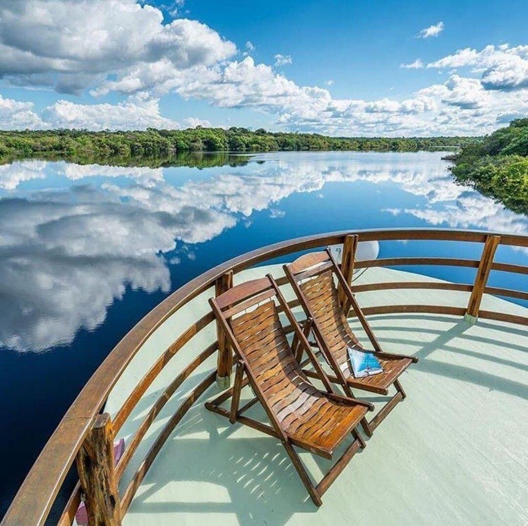 Deux chaises, un fleuve, et un ciel qui ne sait plus où est le haut. Deux chaises longues en bois sur la proue incurvée d'un bateau fluvial lors d'une croisière sur le fleuve Amazone au Brésil, avec des nuages parfaitement reflétés sur les eaux sombres et lisses bordées de forêt