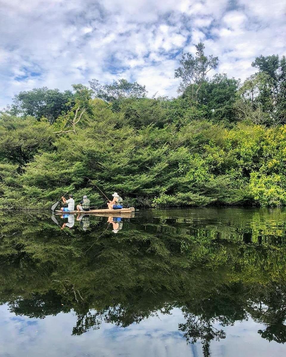The quieter the river, the more the forest has to say. Three travellers in a wooden canoe fishing on a mirror-still river channel during an Amazon River Cruise in Brazil, surrounded by dense tropical forest