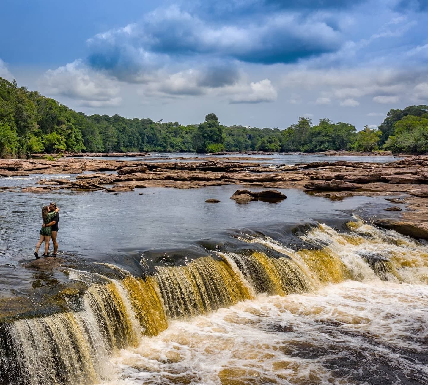 Some moments pick their own backdrop, and the Amazon rarely undersells it. A couple embraces on a rocky ledge at the edge of a wide river waterfall, surrounded by lush rainforest under a dramatic cloudy sky during an Amazon River Cruise Brazil itinerary