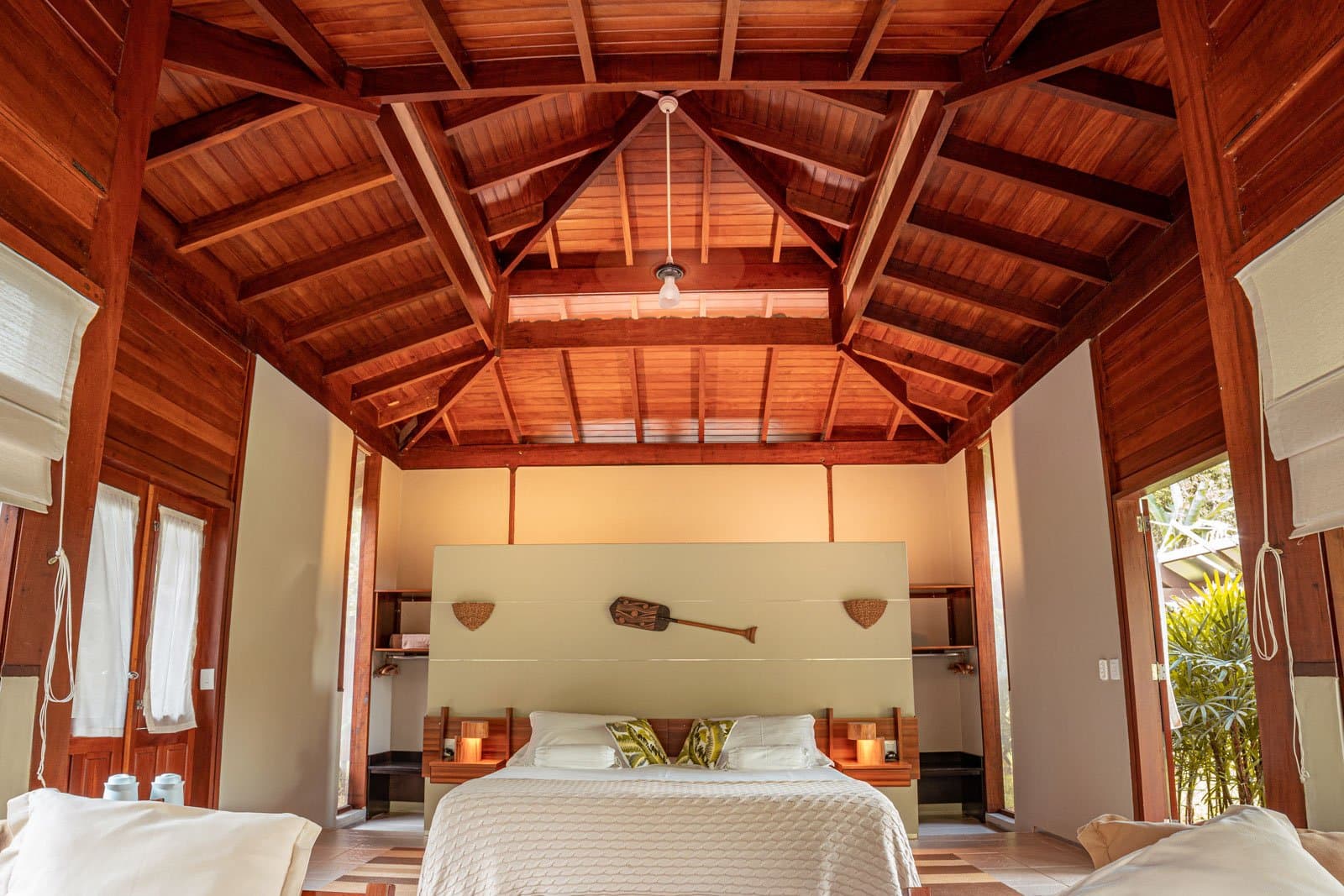 On apprécie le confort quand le réveil sonne à 4h du matin. Photo: Samuel Melim Intérieur spacieux d'un bungalow de lodge de birdwatching en forêt amazonienne, avec un haut plafond en bois à chevrons, un lit double aux draps blancs et coussins à motifs de feuilles, des tables de nuit en bois éclairées à la bougie et une pagaie décorative au-dessus de la tête de lit