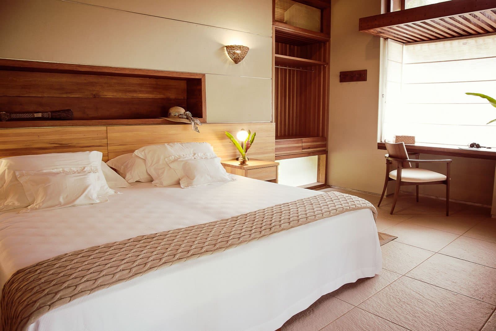 Soft sheets, warm wood, and the forest just outside the window. Photo: | SamuelMelim Photography Warmly lit lodge bedroom with a king-size bed dressed in white linens, wooden headboard with built-in shelving, a bedside lamp with a tropical plant, and a writing desk by a bright window — comfortable base camp for birdwatching in the Amazon rainforest