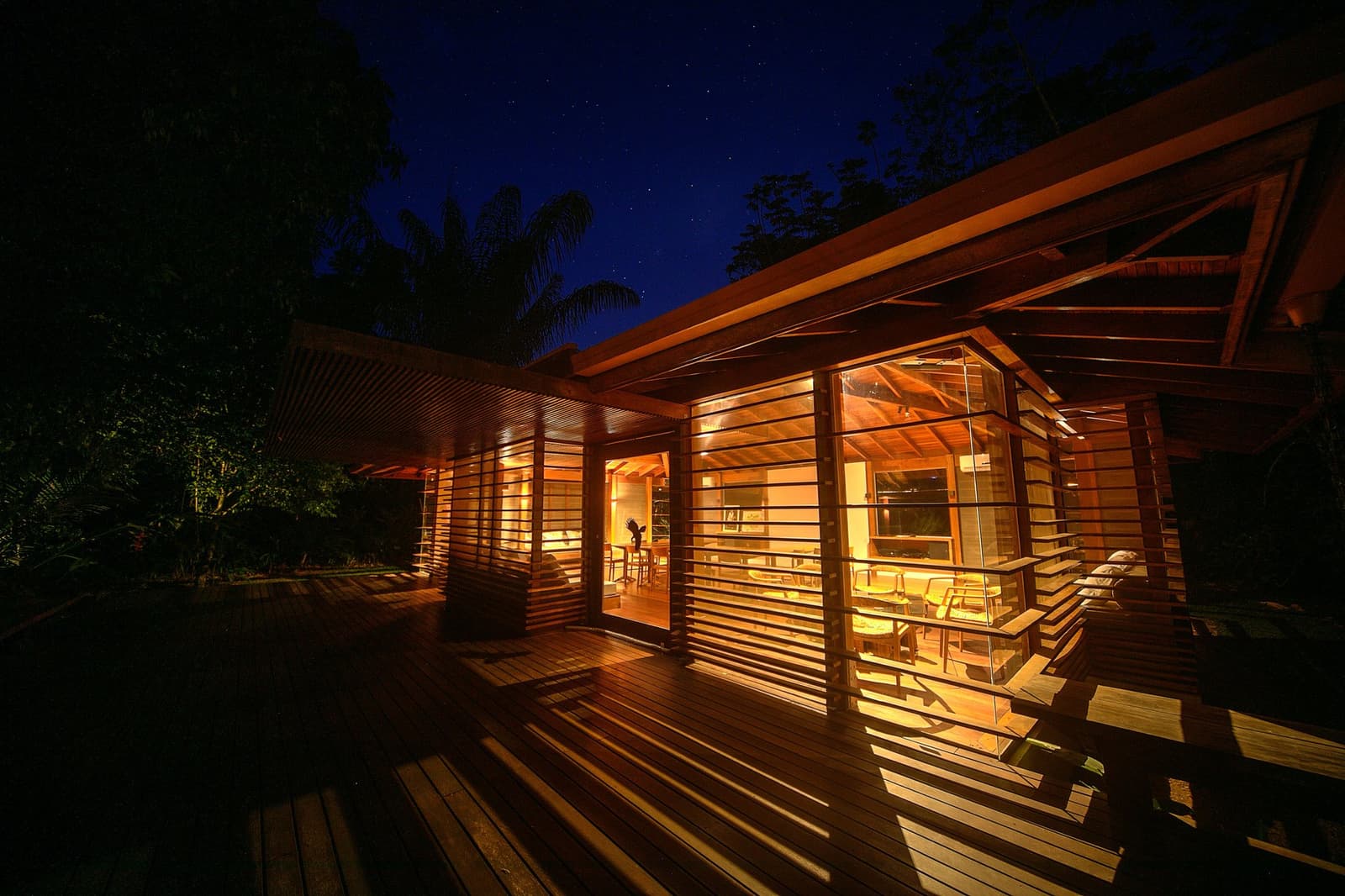 Le dîner est fini, le feu est allumé, et la forêt commence à s'animer. Photo: Samuel Melim Le Cristalino Jungle Lodge illuminé de l'intérieur la nuit, sa façade en bois et verre diffusant une lumière dorée sous un ciel étoilé bleu profond, cerné par la forêt amazonienne dense