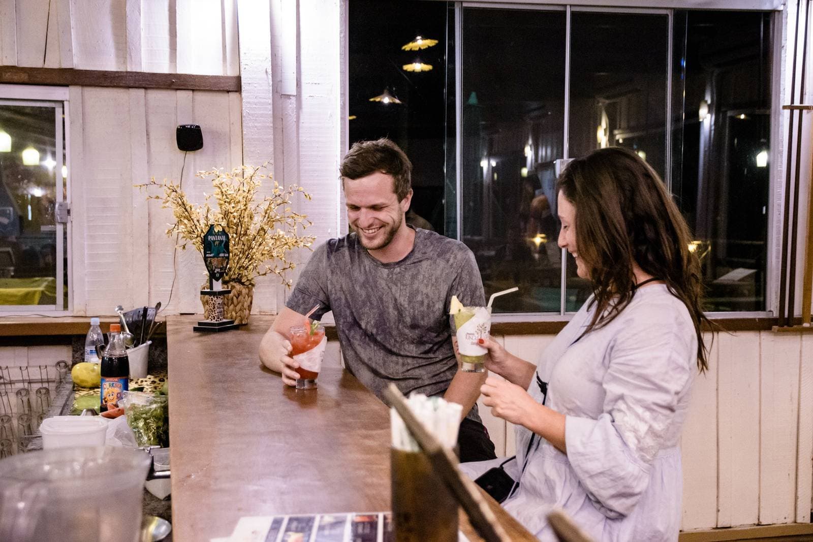 Die besten Pantanal-Geschichten entstehen genau so, Glas in der Hand. Zwei Reisende lachen und halten Cocktails an einer rustikalen Bartheke bei einer günstigen Pantanal-Tour, mit warmem Licht und einem Pantanal-Bierzapfhahn im Hintergrund