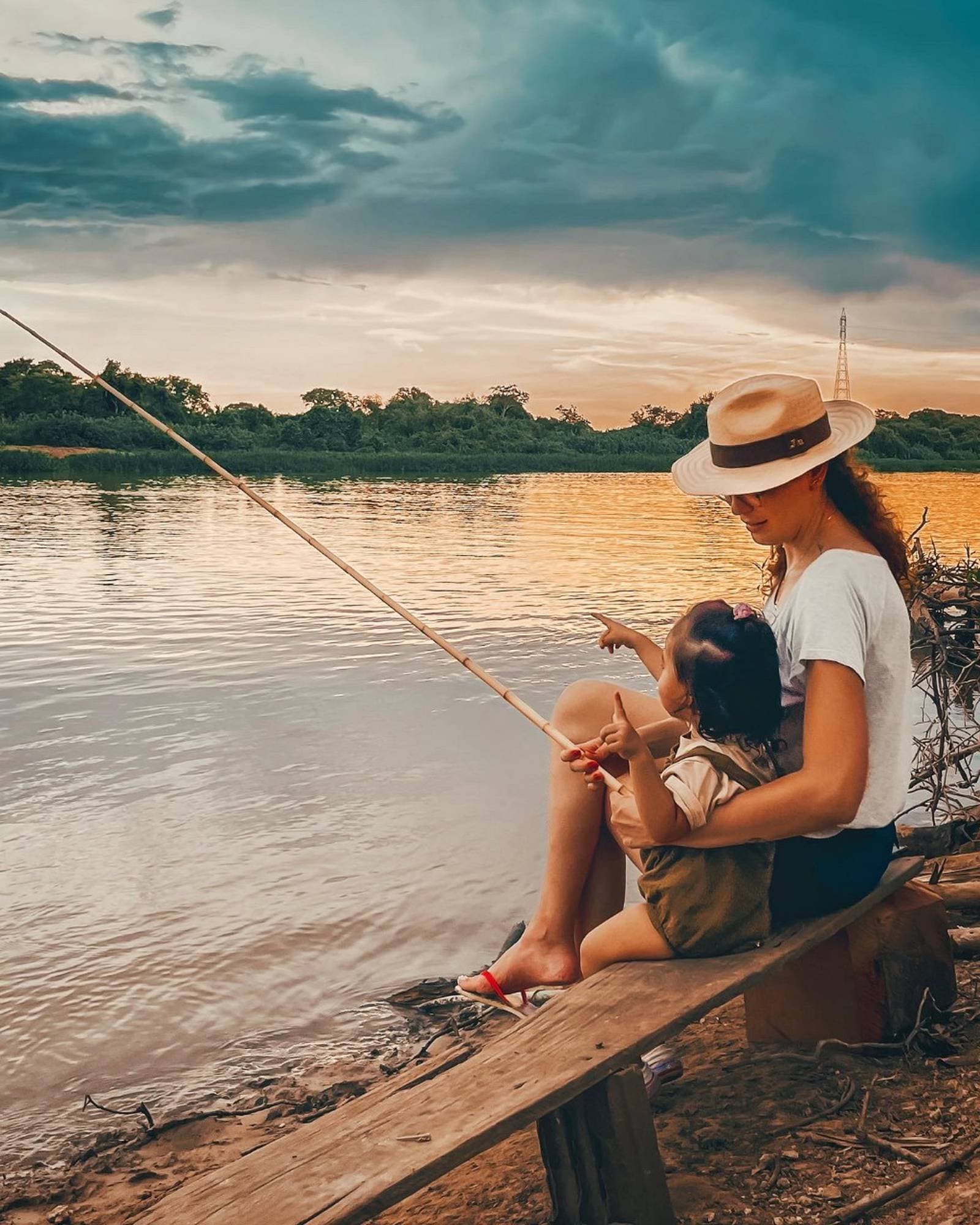 Warten können, der Fluss bringt es bei, das Kind auch. Eine Frau mit Panamahut hält ein Kleinkind auf dem Schoß und angelt mit einer Bambusstange am Flussufer bei Sonnenuntergang, goldenes Licht spiegelt sich auf dem ruhigen Wasser