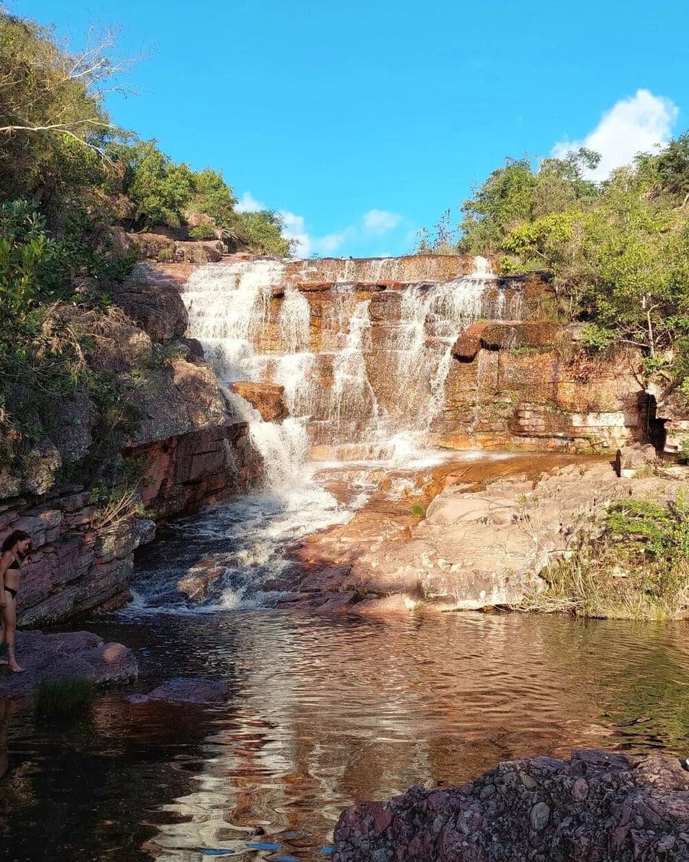 De l'eau froide, de la roche chaude, du temps devant soi. La Chapada fait le reste. Une cascade étagée dévale des dalles de grès aux tons rouille pour rejoindre un bassin naturel limpide lors du Chapada Diamantina Tour, avec une visiteuse au bord de l'eau et une végétation de cerrado sous un ciel bleu