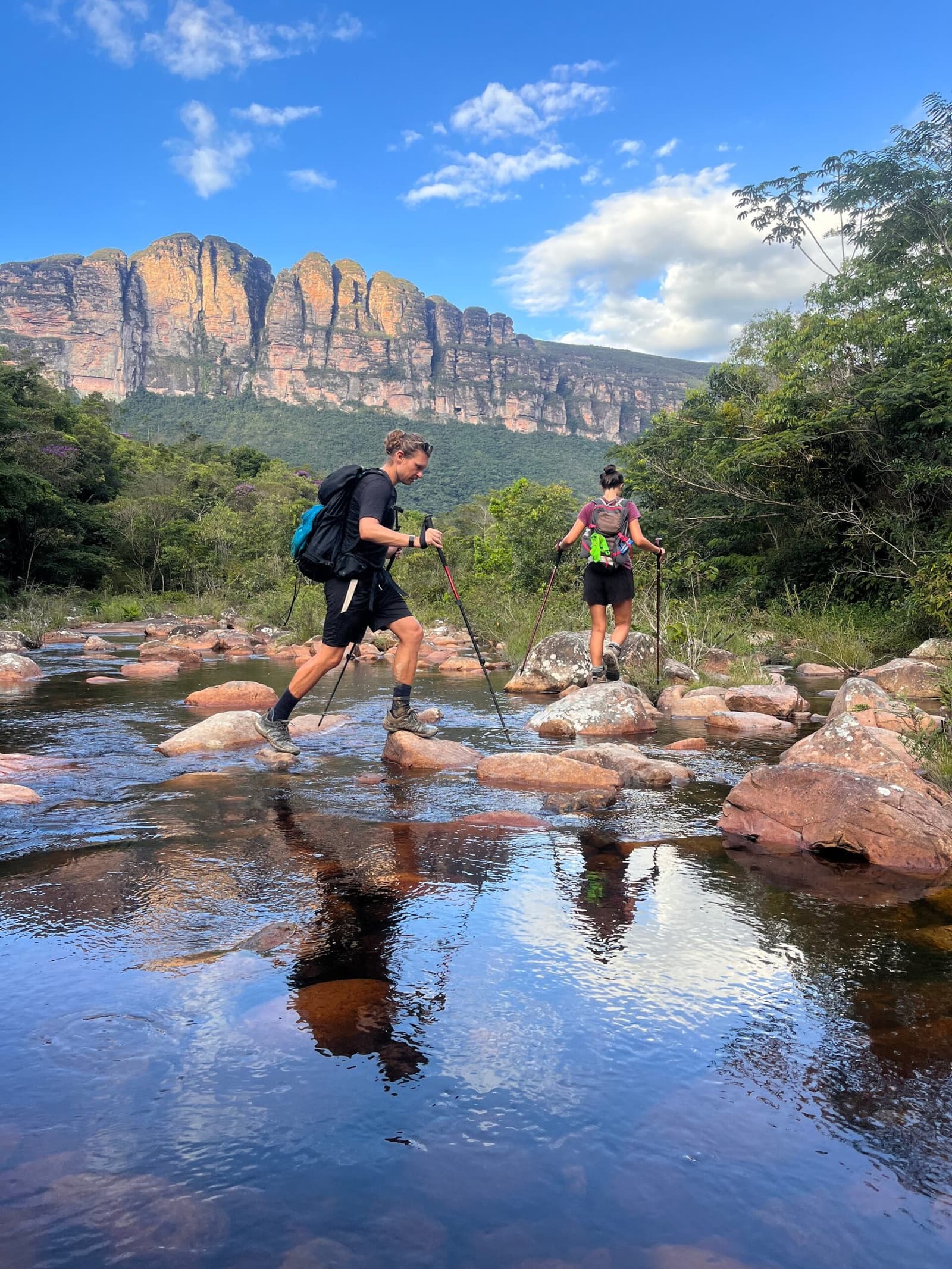 Stein für Stein, die Felsen im Blick. Hier zählt jeder Schritt. Photo: Lucas Neves Zwei Wanderer mit Trekkingstöcken überqueren auf einem Chapada-Diamantina-Trek einen klaren Fluss, indem sie von Stein zu Stein springen, im Hintergrund mächtige Sandsteinfelsen