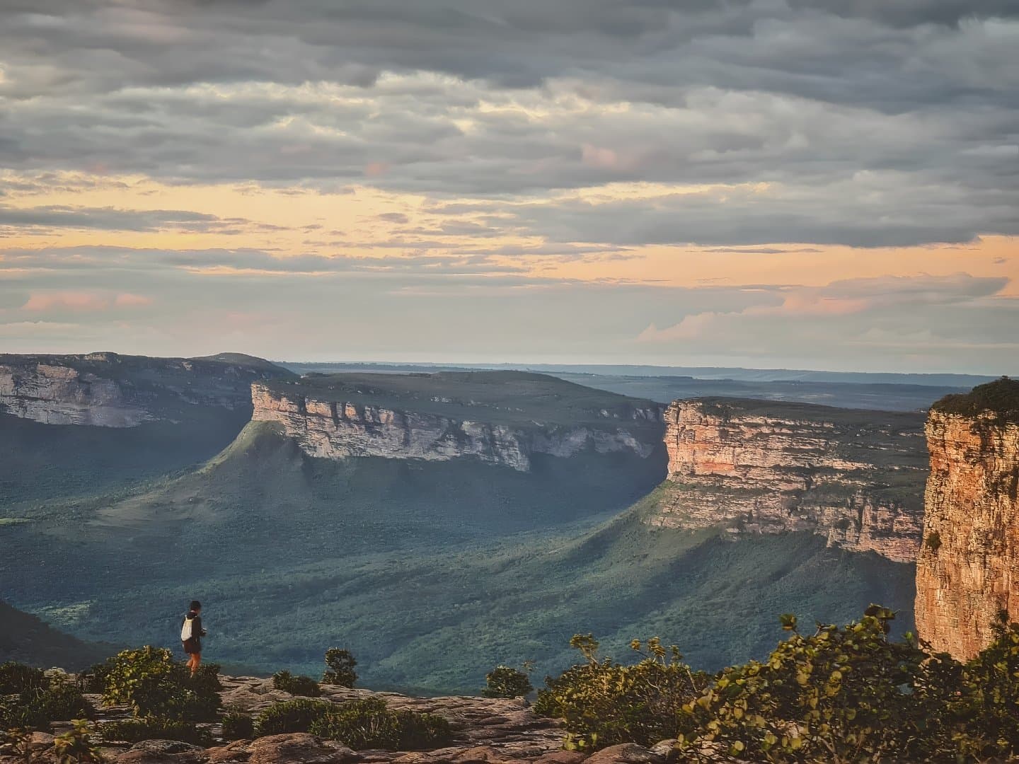 Wenn der Canyon das Wort ergreift, hört man einfach zu. Ein einzelner Wanderer steht auf einem Felsvorsprung mit Blick auf die weiten Sandstein-Tafelberge und grünen Täler der Chapada Diamantina bei Einbruch der Dämmerung, während warmes Licht durch dramatische Wolken bricht