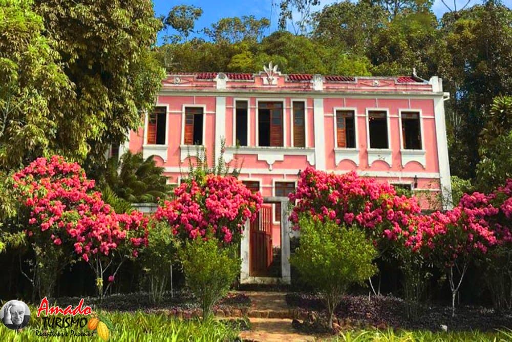 Jardim assim, a gente chega de bike e não quer ir embora. Solar colonial rosa de dois andares da Fazenda Vila Rosa rodeado por buganvílias floridas e Mata Atlântica exuberante na rota de ciclismo do cacau na Bahia
