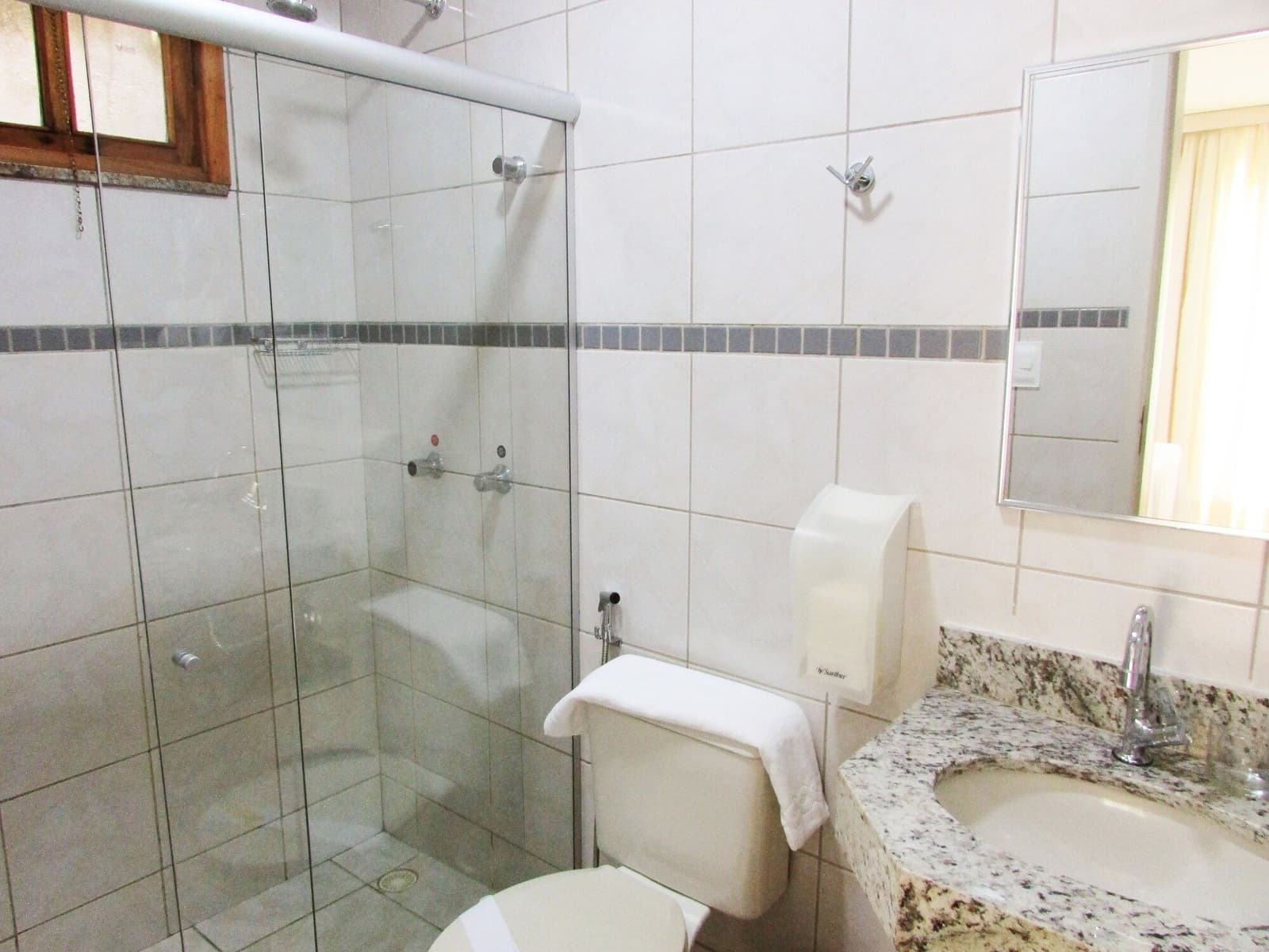 Trinta quilômetros pedalados e o banho quente vira prioridade absoluta. Banheiro privativo limpo em pousada do roteiro de ciclismo na Bahia, com box de vidro, bancada de granito e azulejos brancos com faixa decorativa