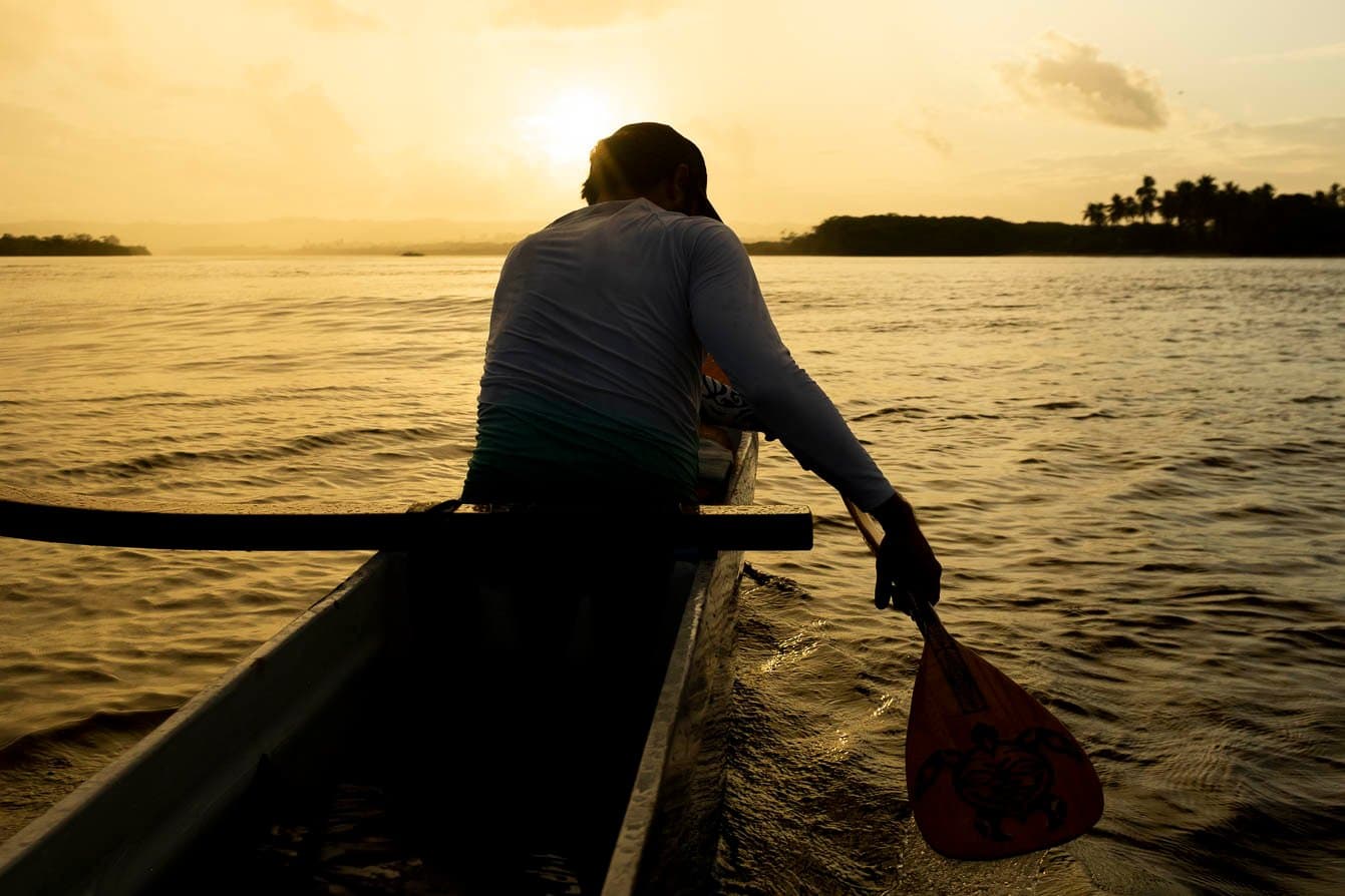 Remo na mão, o rio cuida do resto. Um remador em uma canoa havaiana mergulha seu remo de madeira nas águas calmas do Rio das Contas, em silhueta contra um céu dourado ao pôr do sol com a margem arborizada ao fundo