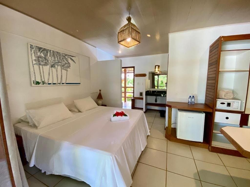 Quarto simples, cama honesta, e o mar esperando lá fora. Quarto de casal claro em uma pousada em Itacaré no roteiro de cicloturismo na Bahia, com roupa de cama branca, luminária de fibra trançada, quadro de palmeiras, frigobar e porta aberta para varanda com jardim