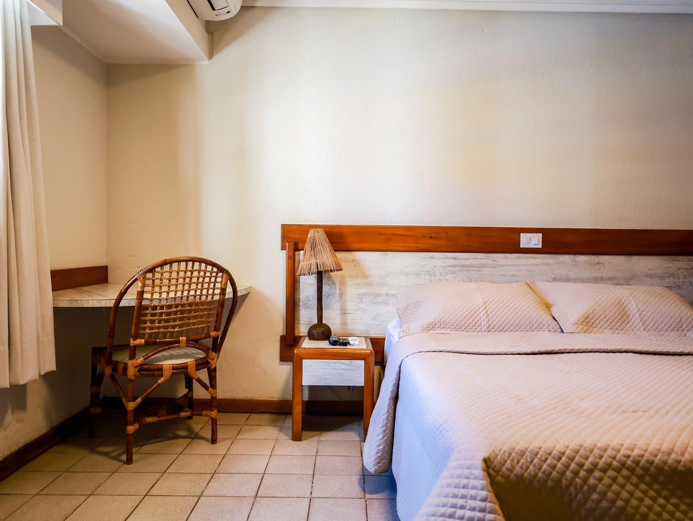 Aconchegante do jeito certo: sem frescura, com tudo que importa. Quarto simples e confortável de pousada em um passeio de ciclismo na Bahia, com cama de casal, colcha clara acolchoada, cadeira de vime, cabeceira de madeira e abajur de bambu trançado