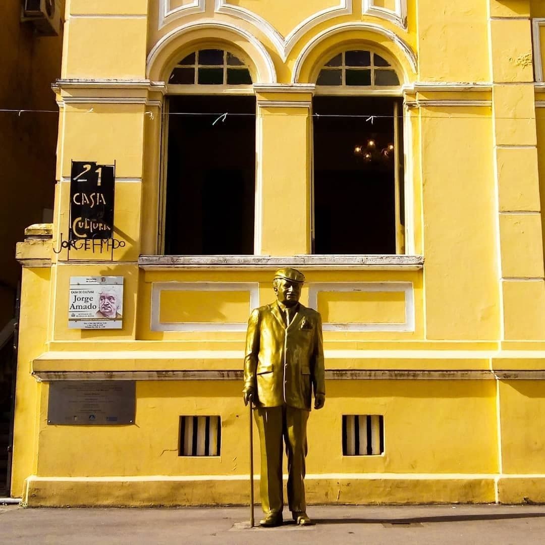 Ilhéus tem Jorge Amado na porta e o mundo inteiro dentro. Estátua dourada de Jorge Amado com bengala em frente à fachada amarela da Casa de Cultura Jorge Amado em Ilhéus, Bahia