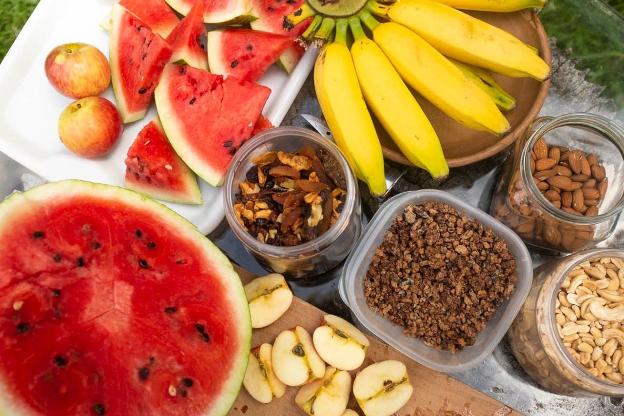 Melancia, castanhas, granola, o carro de apoio não falha. Vista aérea de uma bandeja de lanches para pedalar na Bahia com melancia fatiada, bananas, maçãs, granola, frutas secas, amêndoas e castanhas de caju dispostas ao ar livre