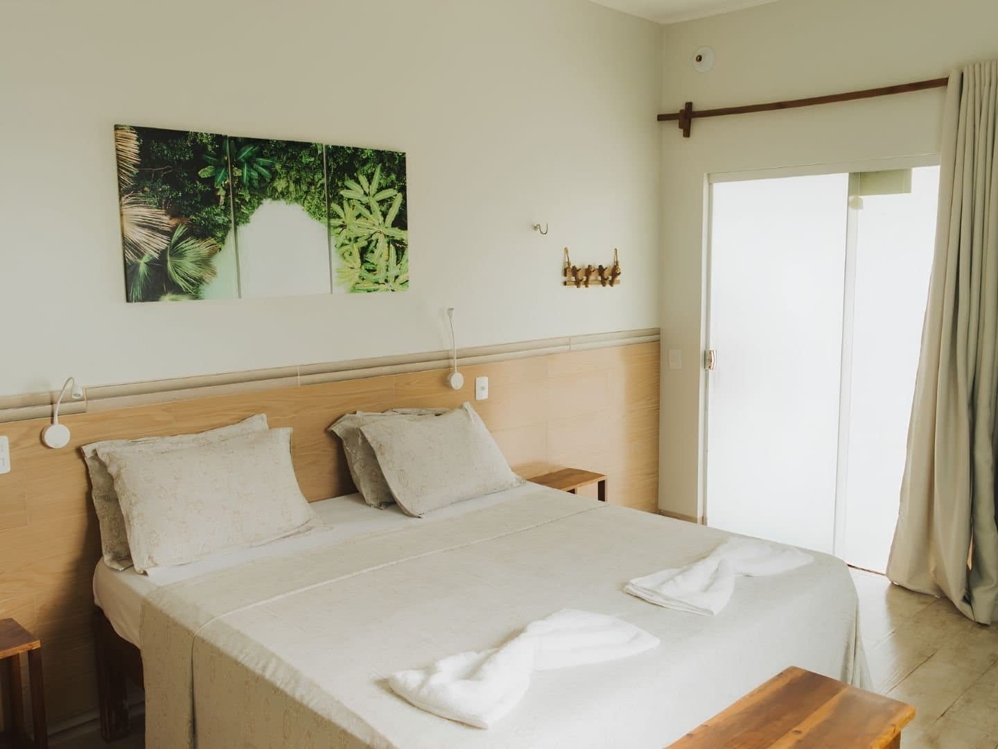 Quarto simples, silêncio de qualidade, sono garantido. Quarto duplo arrumado com roupa de cama branca, cabeceira de madeira, quadros com motivos da natureza e porta de correr para varanda clara, típico das pousadas de uma aventura no Jalapão