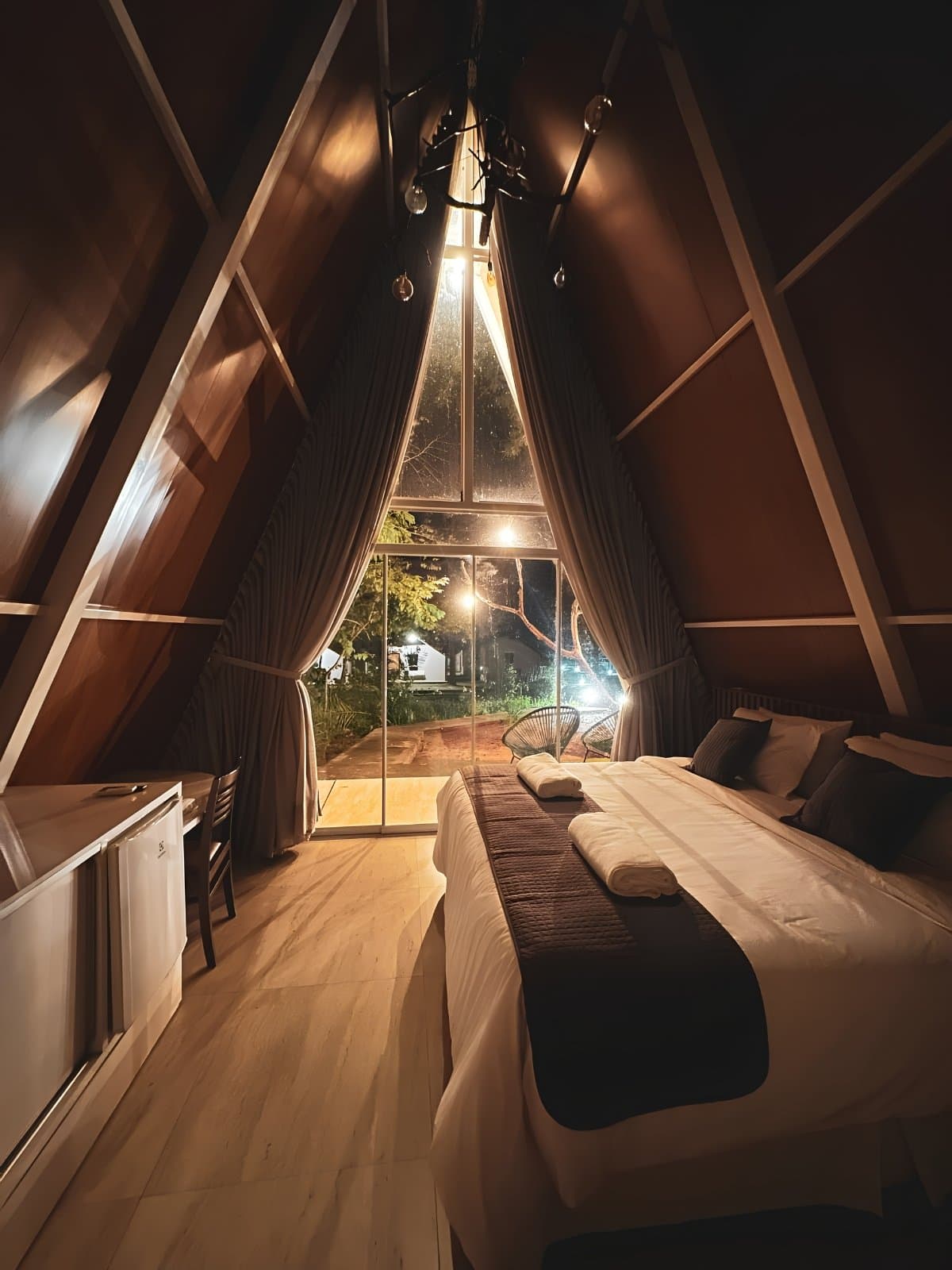 Your legs will thank you for this bed after 15 km on the trail. Interior of an A-frame wooden cabin at night, with a king bed dressed in white linen, warm globe lights hanging from the peaked ceiling, and a floor-to-ceiling triangular window overlooking a lit garden — comfortable lodging on a Jalapão trekking itinerary