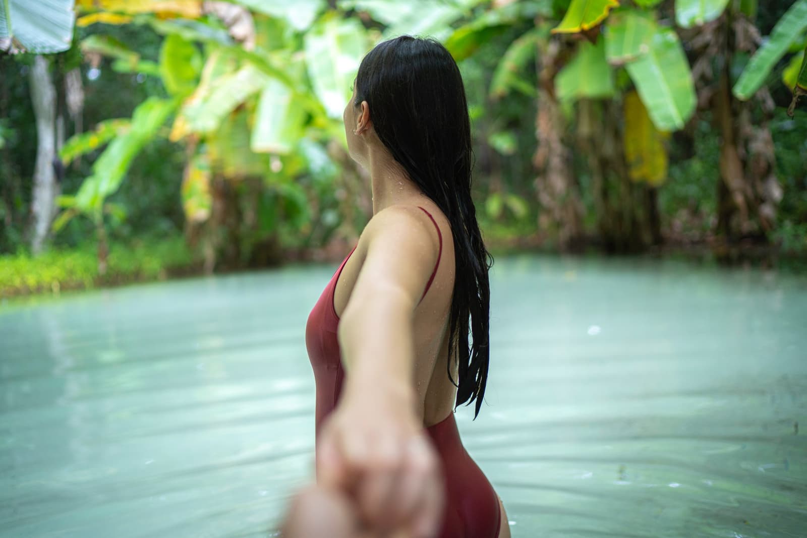 Cool water, no bottom to touch, just the pull of a fervedouro doing its thing. Photo: FG Trade A woman in a red swimsuit wading waist-deep in a clear, milky-blue natural spring surrounded by dense tropical foliage during a Jalapão trekking immersion, reaching her hand back toward the camera