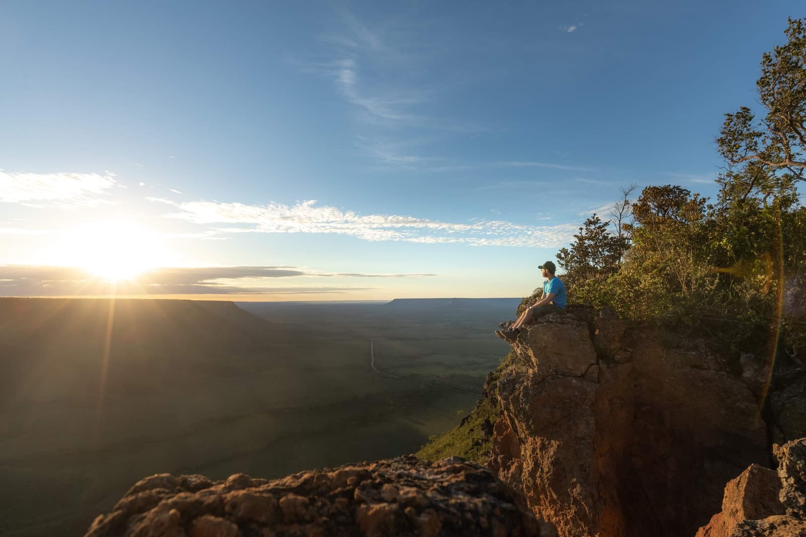 Sit here long enough and the whole park sorts itself out below you. Photo: Alexandre Socci A hiker in a blue shirt sits on the edge of a sandstone escarpment during Jalapão trekking, watching the sun set low over a vast cerrado plateau stretching to the horizon