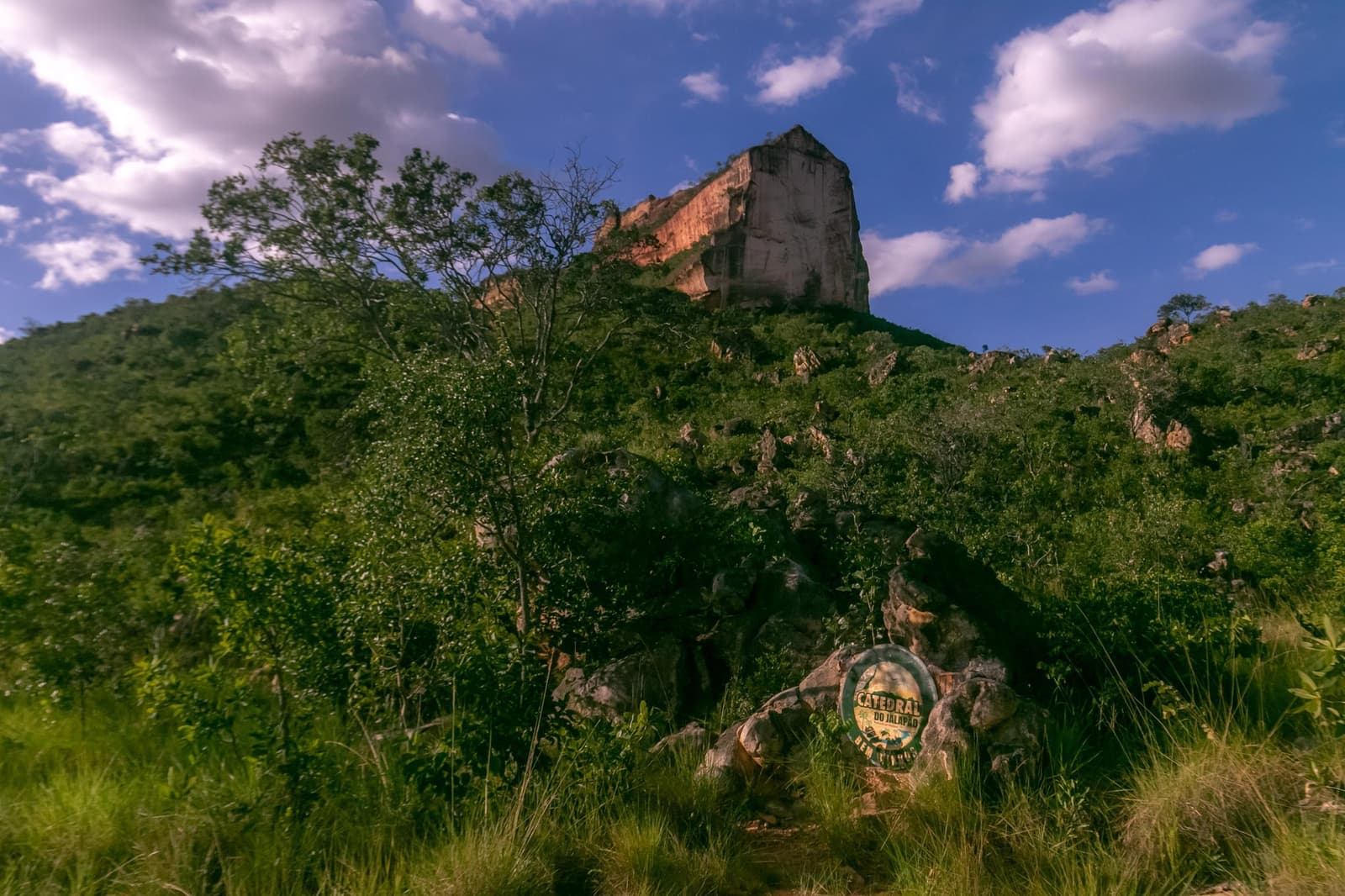 That sign on the rock is the first hint of what's waiting at the top. Photo: Alexandre Socci Low-angle view of the massive sandstone pillar of Catedral do Jalapão rising above a green cerrado hillside, with a circular trail marker sign visible among the rocks in the foreground and a blue sky with scattered clouds above, on a jalapão trip