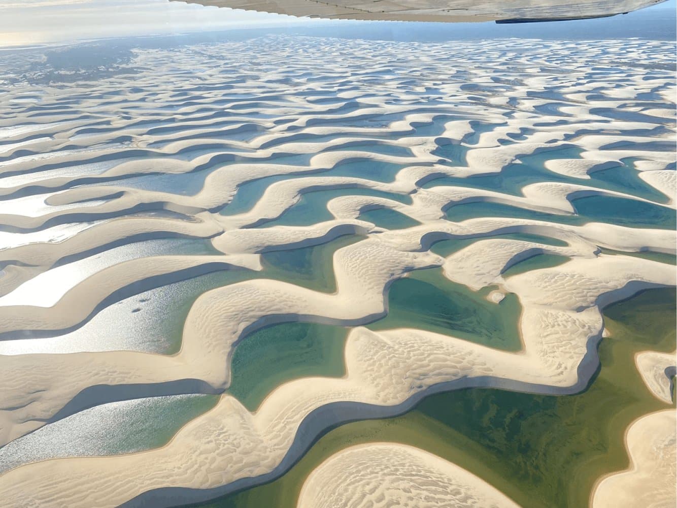 Daqui de cima, fica difícil acreditar que isso é real, e não é pintura. Vista aérea a partir de uma pequena aeronave sobre as dunas de areia branca e lagoas turquesa e esmeralda dos Lençóis Maranhenses estendendo-se até o horizonte
