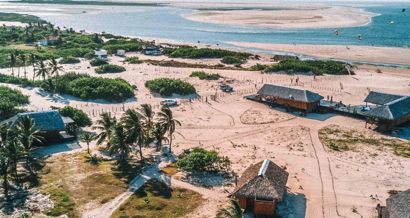 Poucas casas, muito vento e uma imensidão de areia ao redor. Vista aérea de cabanas com telhado de palha, coqueiros e trilhas de areia no vilarejo rústico de Atins, com papagaios de kite visíveis nas águas azuis ao fundo