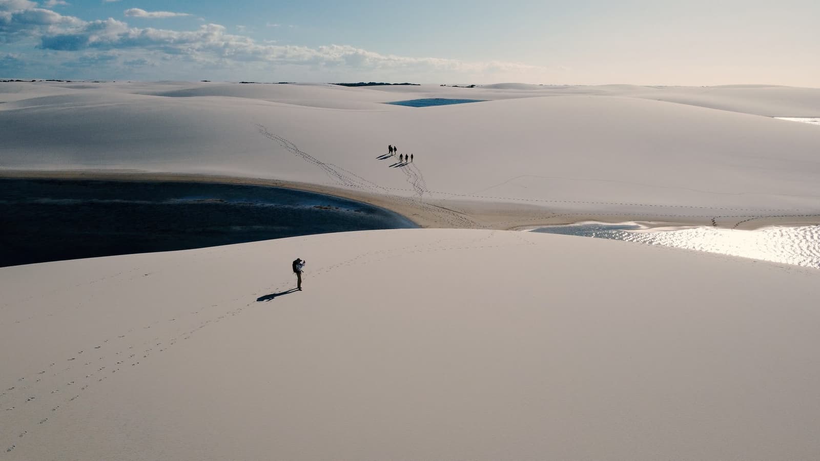 As dunas engolem qualquer noção de escala, e a gente só percebe isso quando já está no meio delas. Vista aérea de caminhantes deixando pegadas nas imensas dunas brancas do roteiro completo dos Lençóis Maranhenses, com lagoas de água escura aninhadas entre as cristas de areia