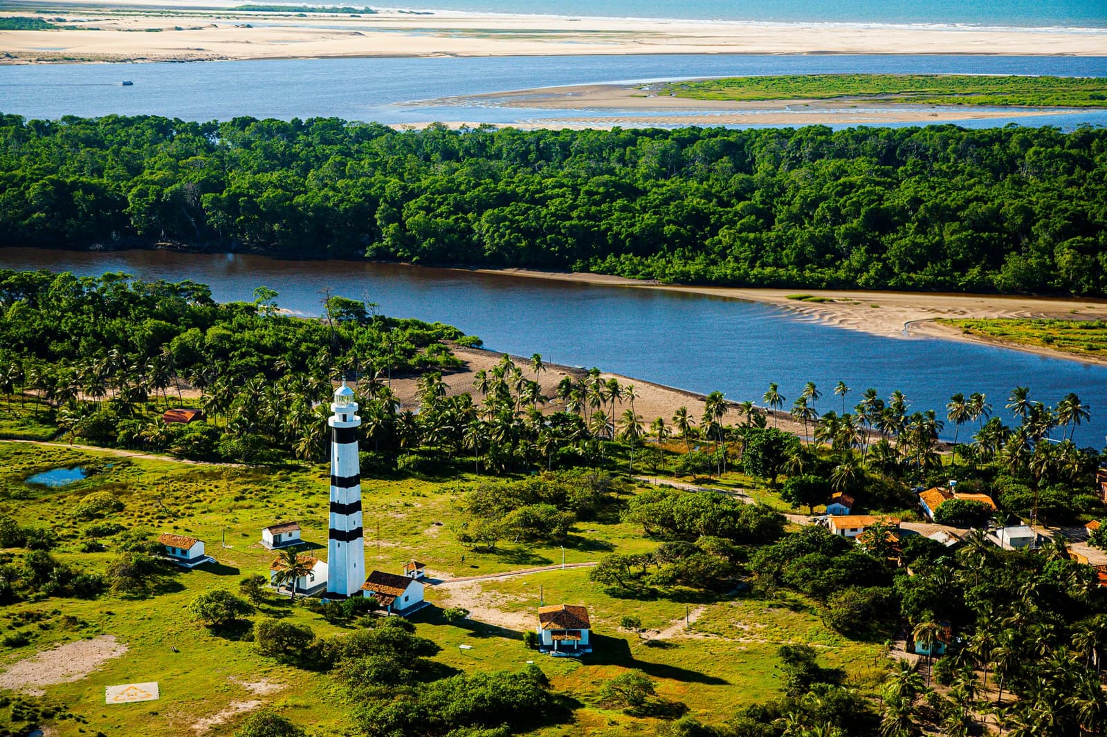 O farol conhece essa paisagem de cor, muito antes de qualquer turista chegar. Vista aérea do farol de Mandacaru, com listras pretas e brancas, cercado por palmeiras e pequenas construções, com o rio Preguiças, floresta densa e dunas de areia branca ao fundo — ponto de destaque do roteiro completo pelos Lençóis Maranhenses